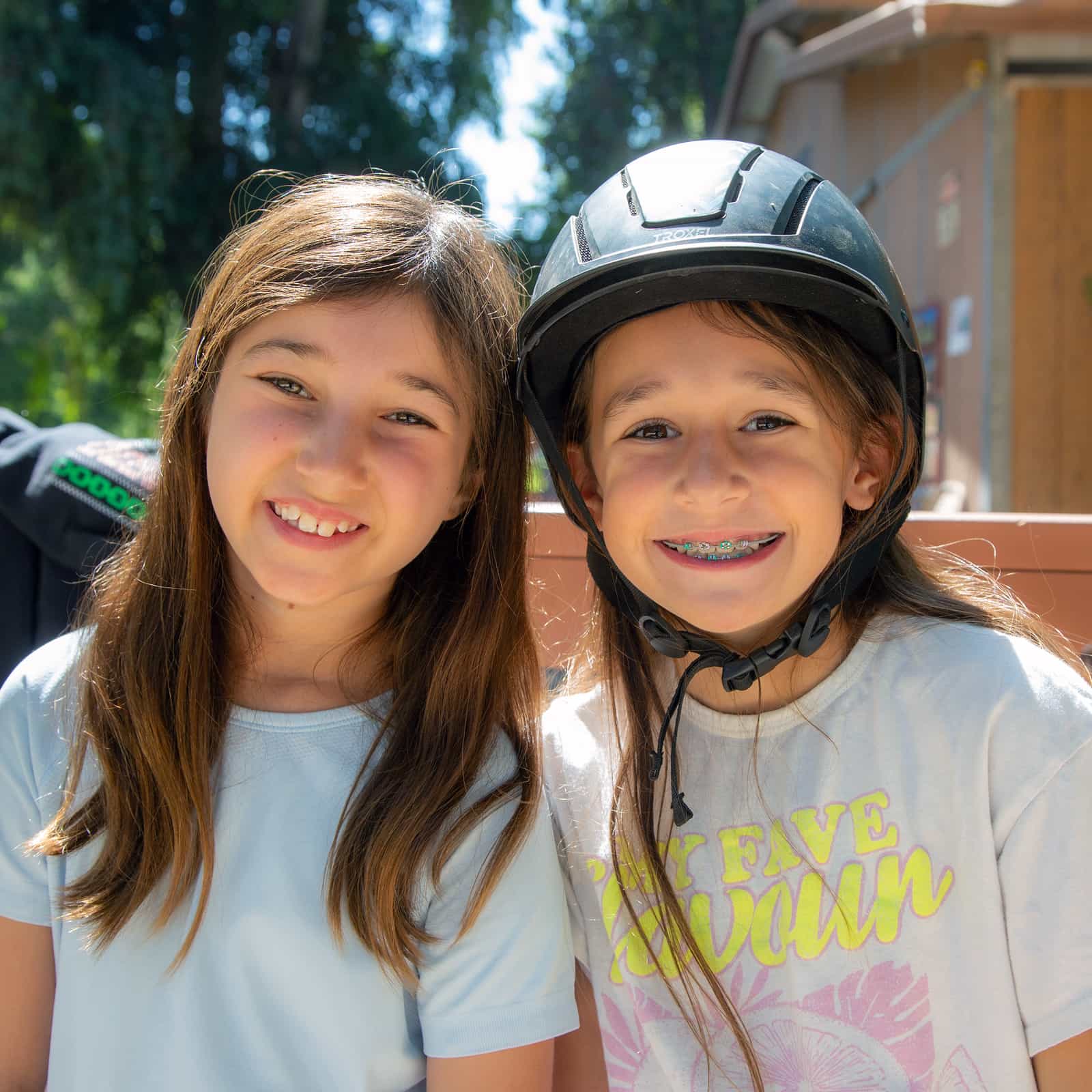 Two smiling campers sitting outdoors at Camp Alonim, with trees and a cabin in the background. One camper wears a helmet and braces, and both appear relaxed and happy in the sunshine.