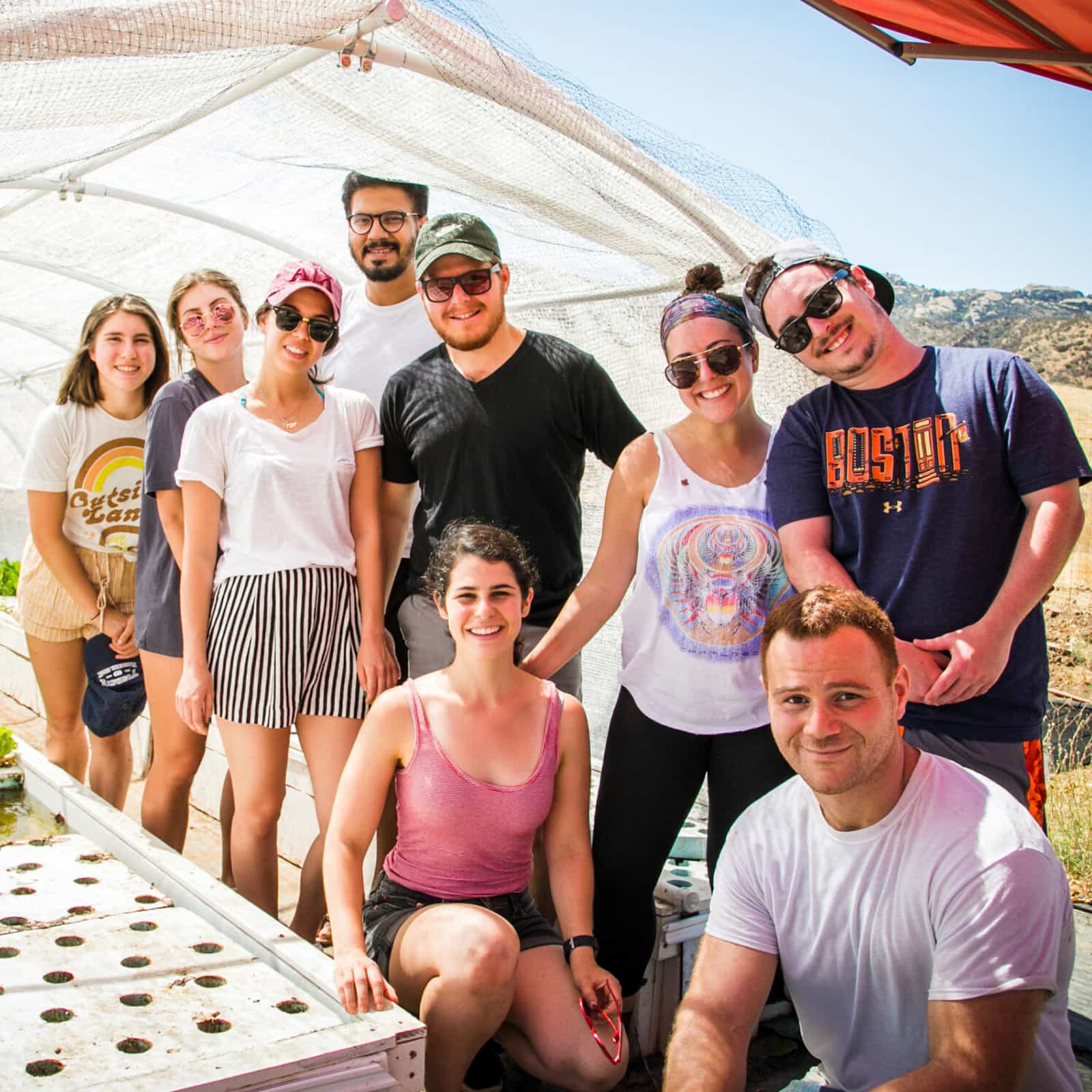 Group of Ziering Brandeis Camp Institute participants smiling together outdoors under a shade canopy, standing beside a garden growing setup. The image represents community-building and leadership experiences for Jewish young adults.