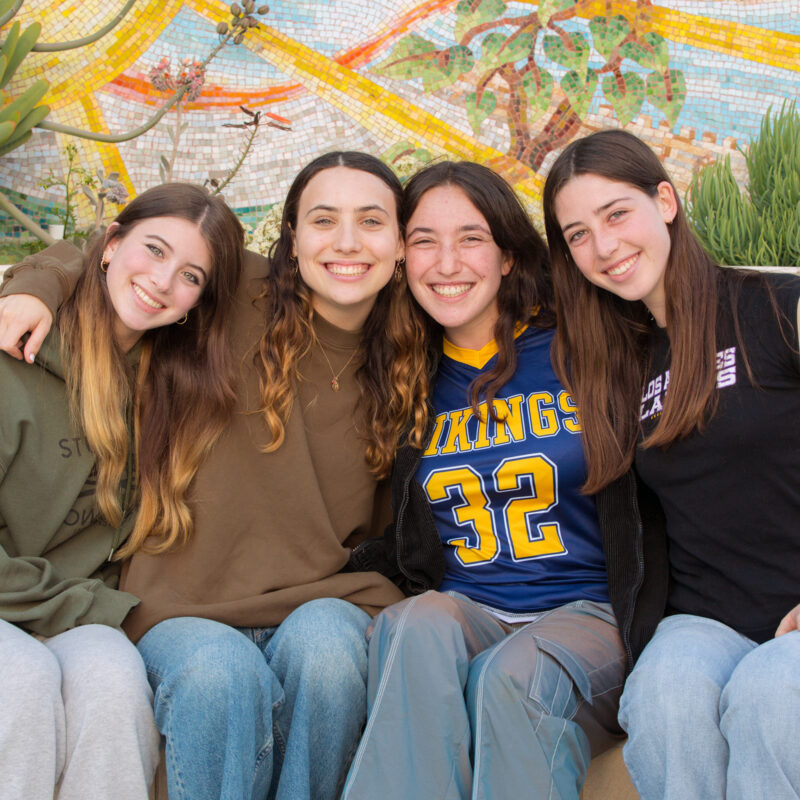 Four teens smiling together in front of a colorful mural, representing participants in a Jewish Learning Experience program.