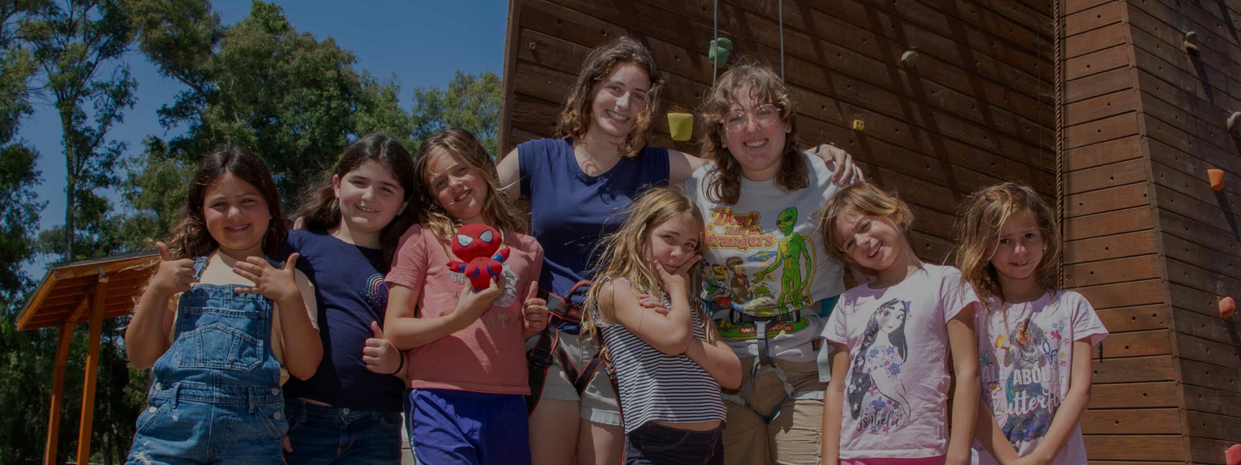 Camp counselors and campers smiling together in front of a rock climbing wall at Camp Alonim.