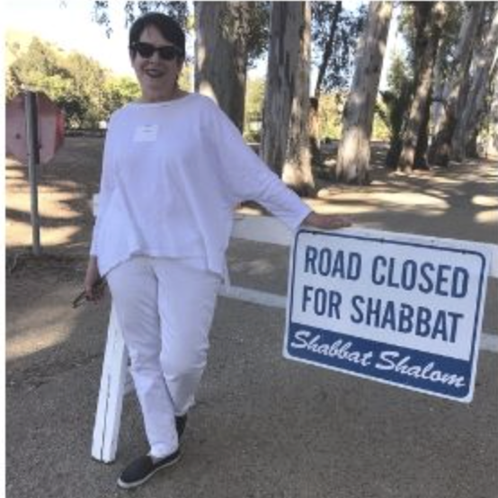 A photo of a woman out in a forest, dressed in all white, in front of a "road closed for Shabbat" sign.
