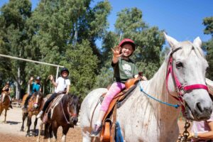 A young girl on a horse smiling and waving at the camera with other kids on horseback behind her.
