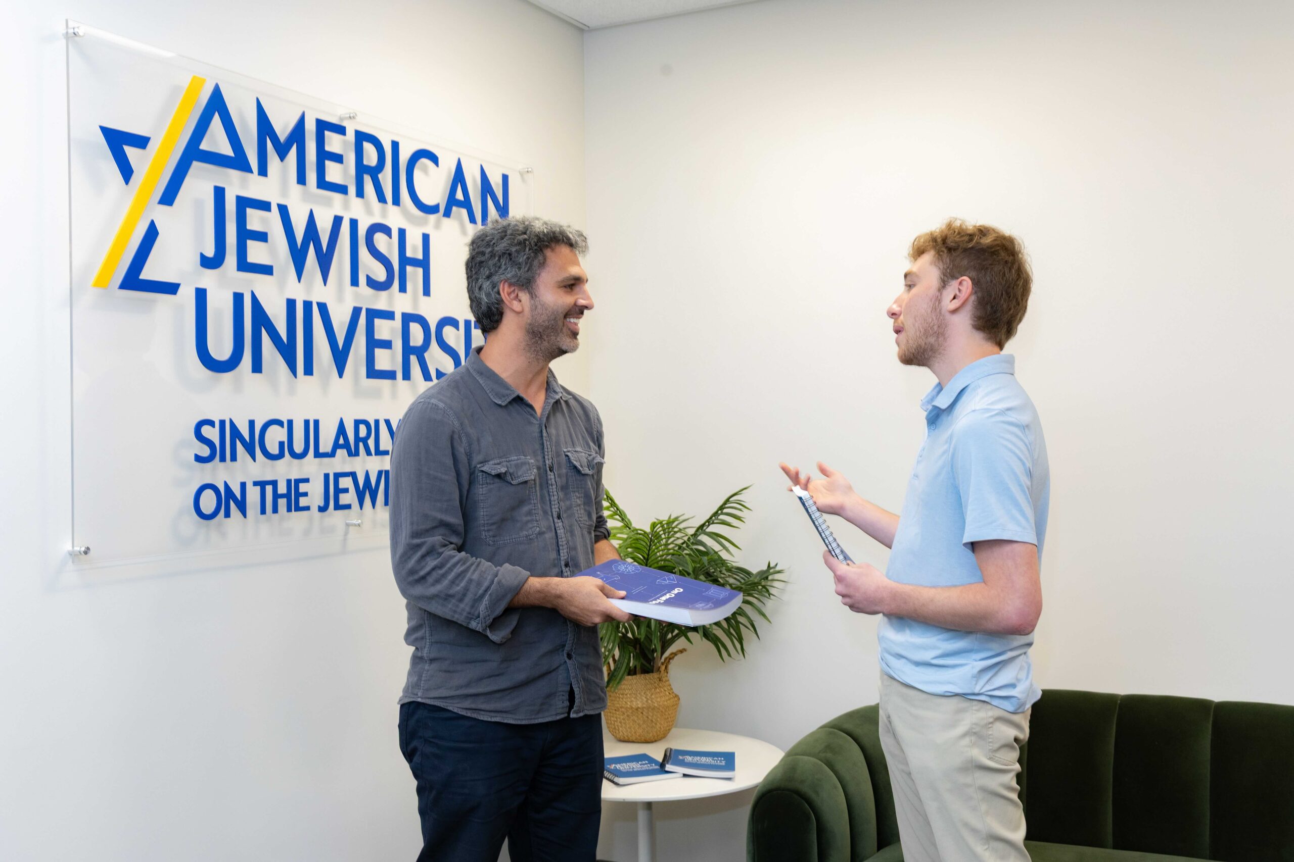 Two men chatting with each other and smiling in front of the AJU logo on a wall.