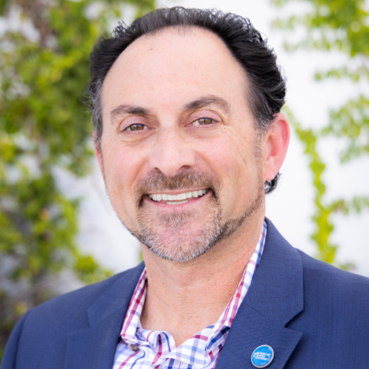 Headshot of Jay Greenlinger with checkered collar and blue jacket with AJU pin
