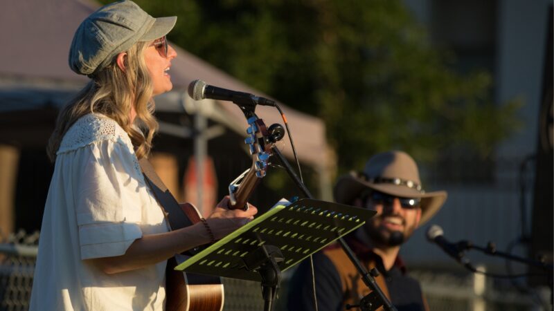 Woman playing guitar and signing into microphone with a smiling man behind her playing guitar