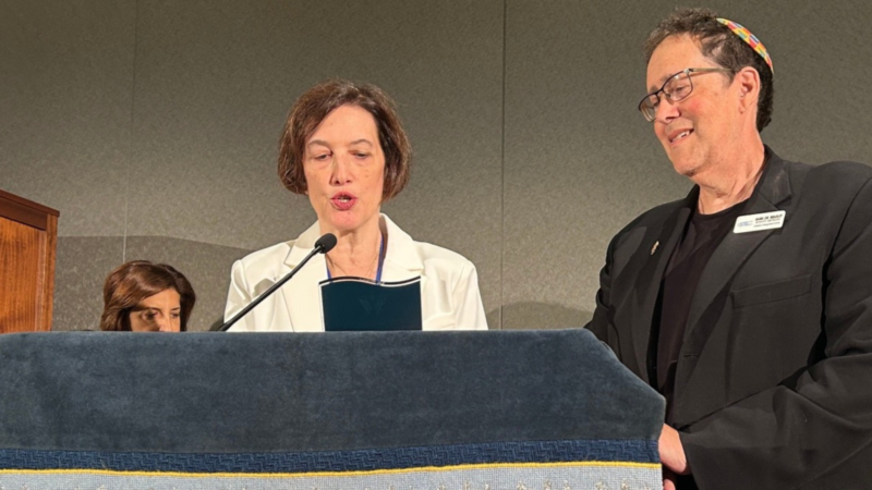Bradley Artson smiling and standing next to a podium with a woman speaking