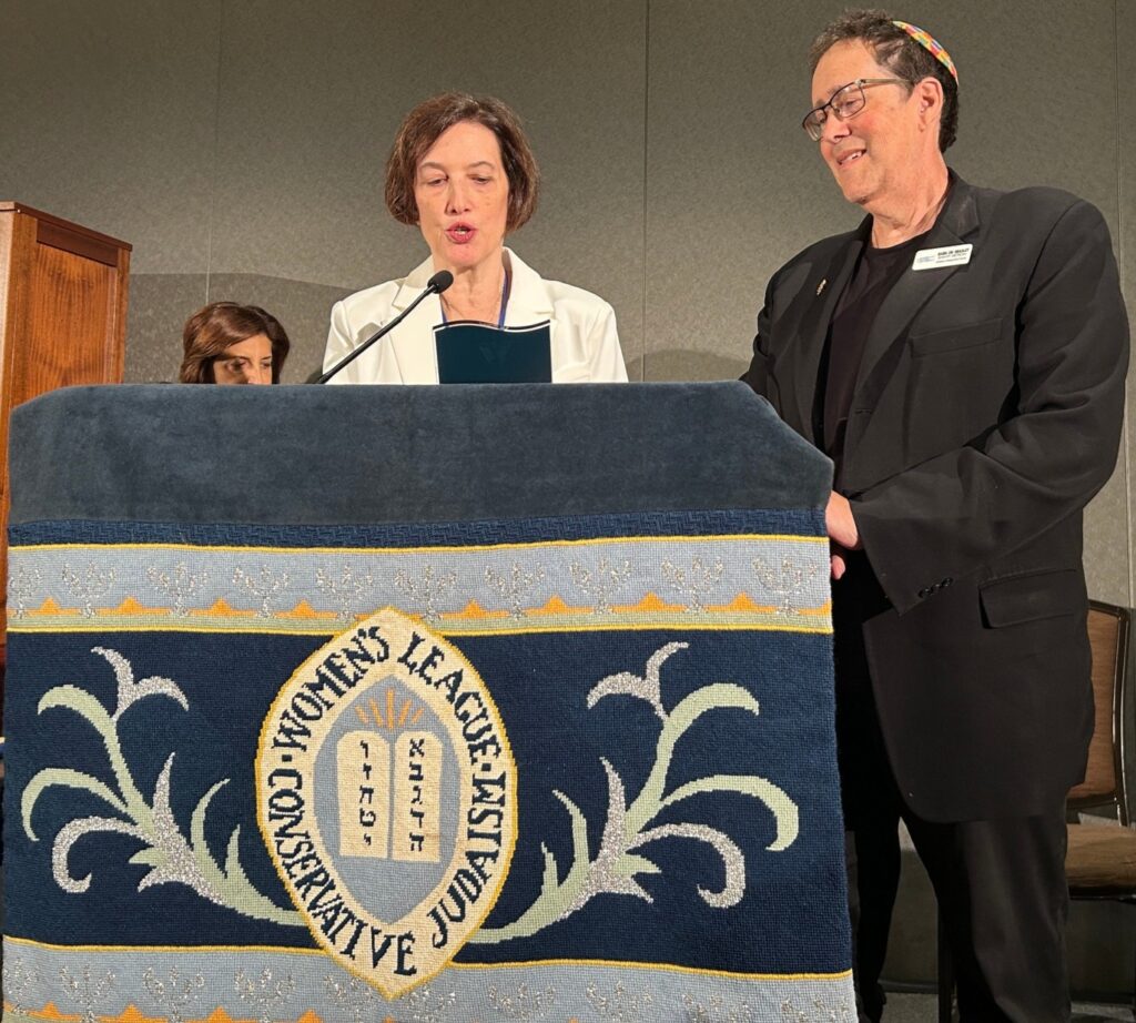 A woman in a white coat from Women's League standing at a podium with the Womens League crest, Rabbi Artson in a black suit standing next to the podium