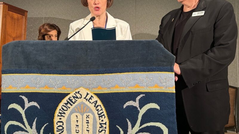 A woman in a white coat from Women's League standing at a podium with the Womens League crest, Rabbi Artson in a black suit standing next to the podium