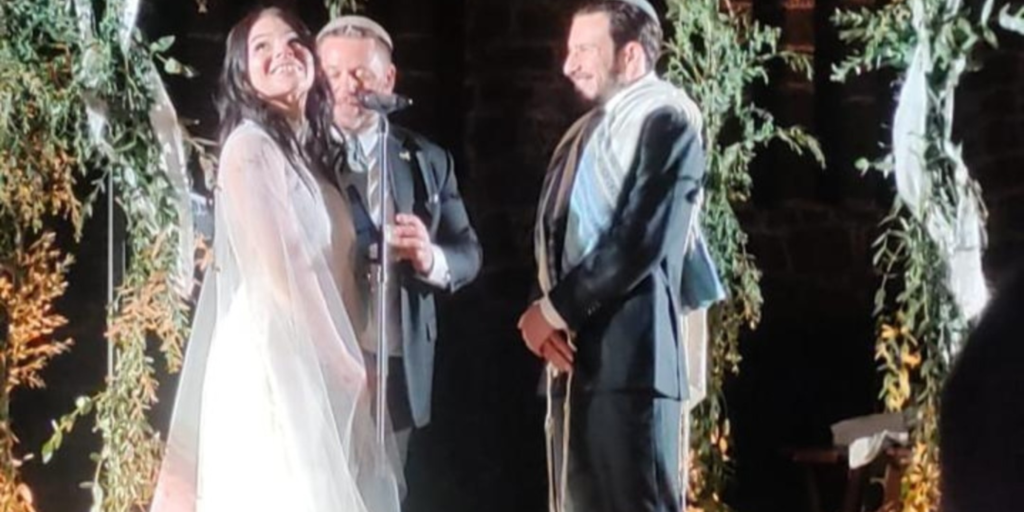 Eduardo in a suit and talis and Claudia in a long white wedding dress and veil under the chuppah with a rabbi in a suit
