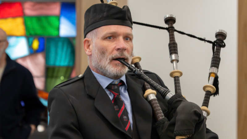 Kevin McIntyre in a black suit and black kippah playing the bagpipes in a synagogue with stained glass windows