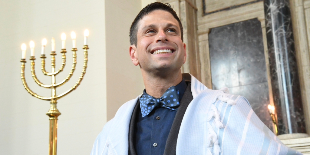 Rabbi Burg smiling in a blue shirt and blue pola dot bowtie with and a white tallit, standing next to a menorah