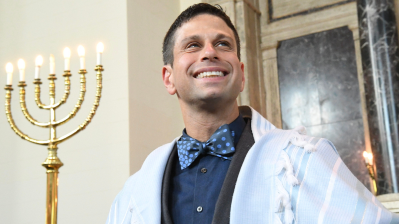 Rabbi Burg smiling in a blue shirt and blue pola dot bowtie with and a white tallit, standing next to a menorah