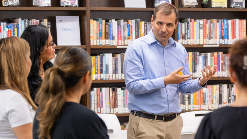 A man in a blue button down shirt and khakis stands in front of a library shelf filled with books and lectures to four students