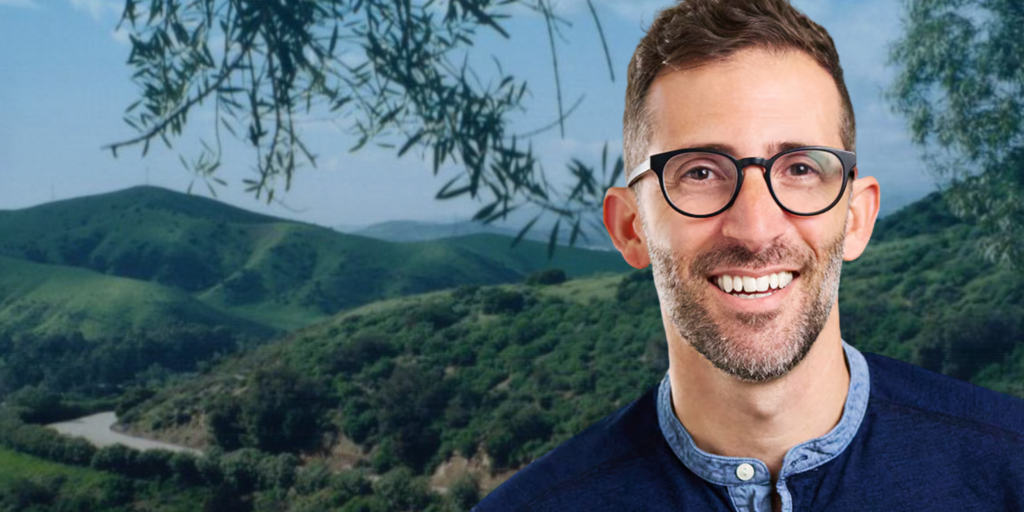 Ari Bloom in black glasses and a blue shirt against the BBC backdrop