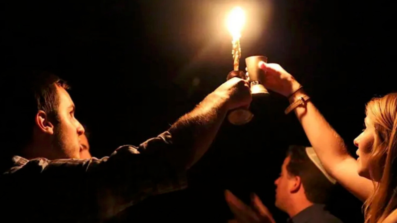 A man holding up a lit havdalah candle and a woman holding up a kiddush cup in the darkness