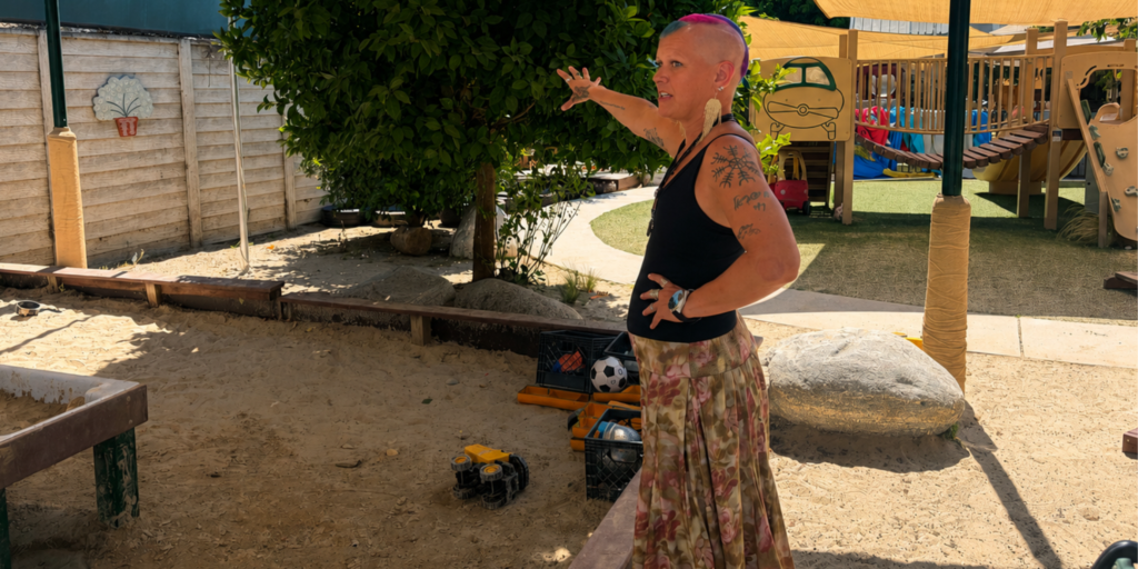 Chana Blugrind with a rainbow buzzcut in a black tank top and flowery skirt stands in a children's playground