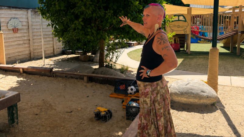 Chana Blugrind with a rainbow buzzcut in a black tank top and flowery skirt stands in a children's playground