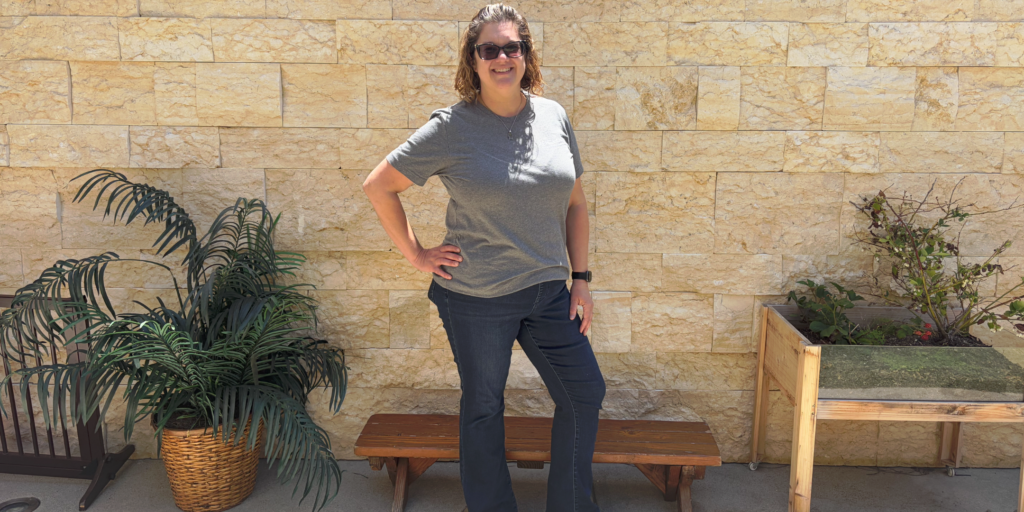 Katie Kang is in jeans and a gray t shirt and sunglasses and is standing in front of a sandstone wall with plants