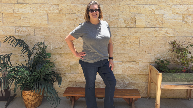Katie Kang is in jeans and a gray t shirt and sunglasses and is standing in front of a sandstone wall with plants