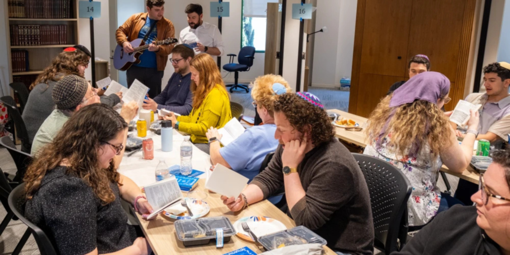 Twelve people sit around two long tables studying from books while two men at the front, one with a guitar, lead them in study