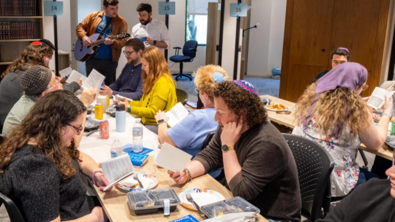 Twelve people sit around two long tables studying from books while two men at the front, one with a guitar, lead them in study
