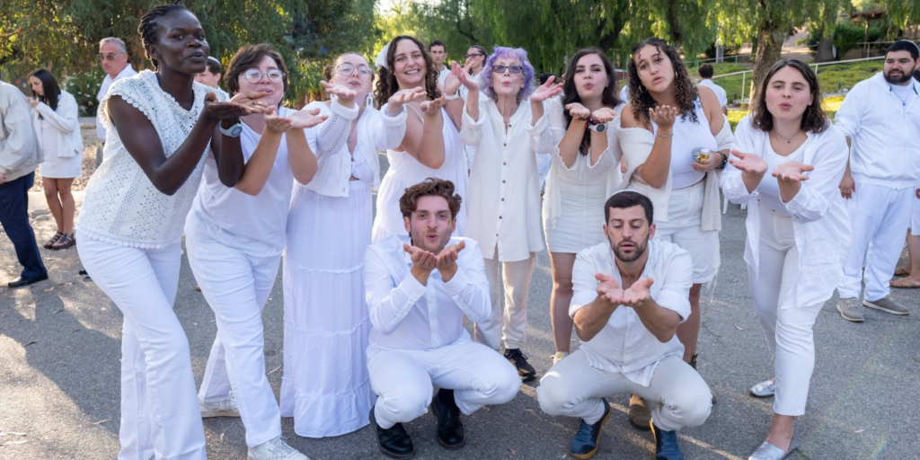 10 people in all white blow a kiss at the camera while other people in white look on in the background