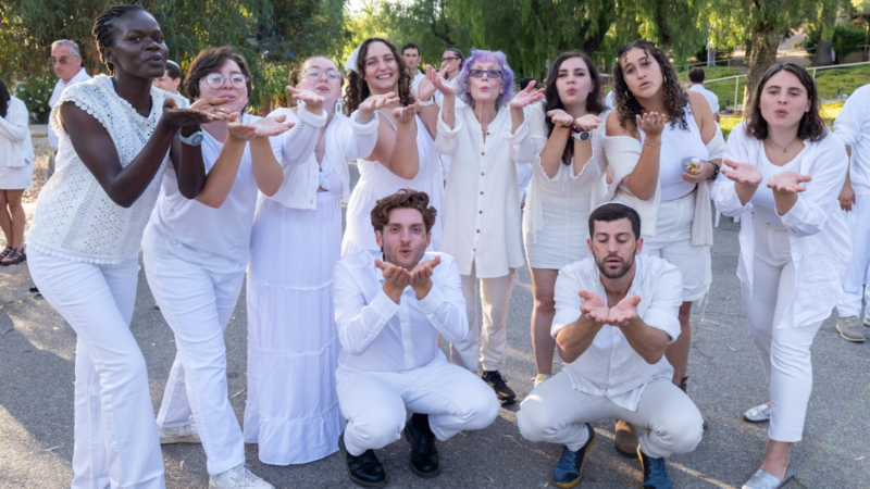 10 people in all white blow a kiss at the camera while other people in white look on in the background