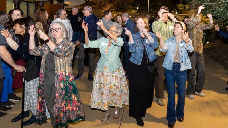 A multigenerational group of attendees smiles, dances, and raises their hands during an outdoor evening celebration of the Ziering Brandeis Camp Institute’s 85th anniversary.