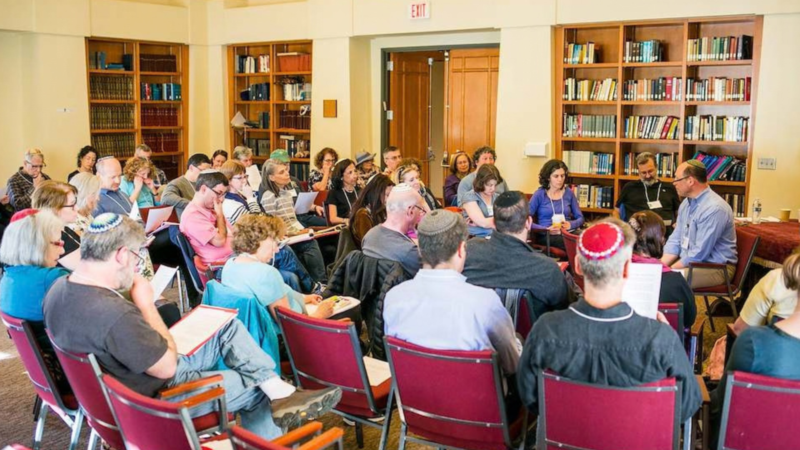 A large room lined with books and filled with people in yarmulkes in red chairs studying a text