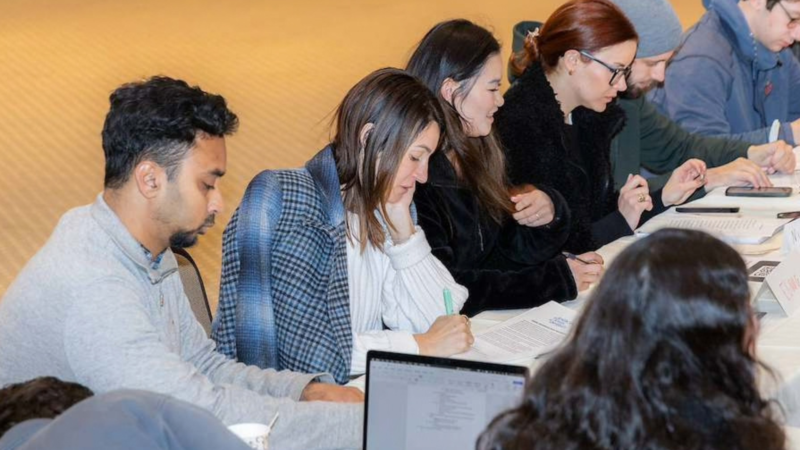 Six people sit at a long table and study Jewish texts in hard copy and on a laptop computer