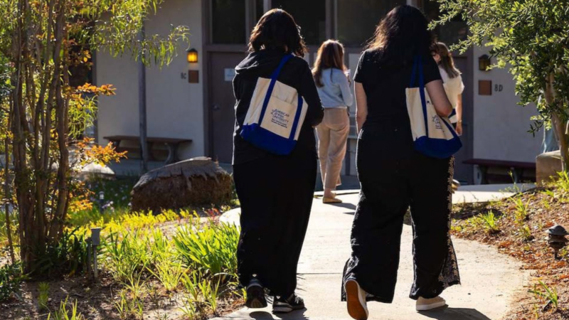 Four women walk outside, two holding American Jewish University tote bags with blue straps