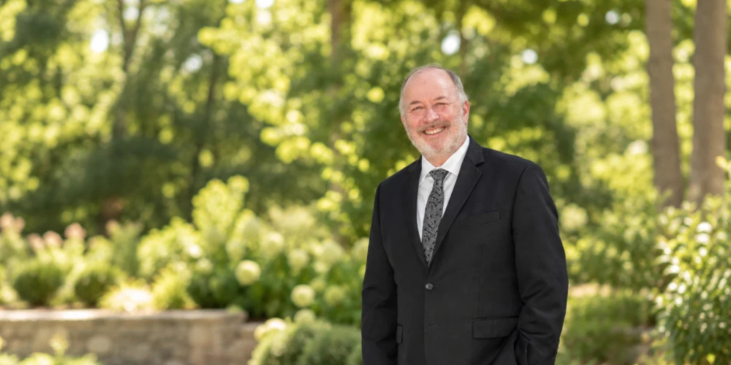 Rabbi Stuart Vogel smiles in a black suit, white shirt, and gray tie in front of a grove of trees