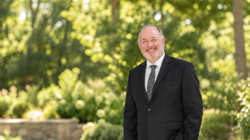Rabbi Stuart Vogel smiles in a black suit, white shirt, and gray tie in front of a grove of trees