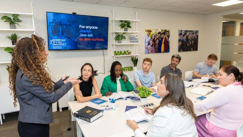 A woman in a gray jacket speaks to 7 adult students witting a table covered in Jewish texts