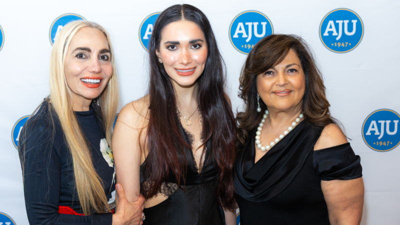Three women in black evening gowns standing in front of a step and repeat with the aju logo on it