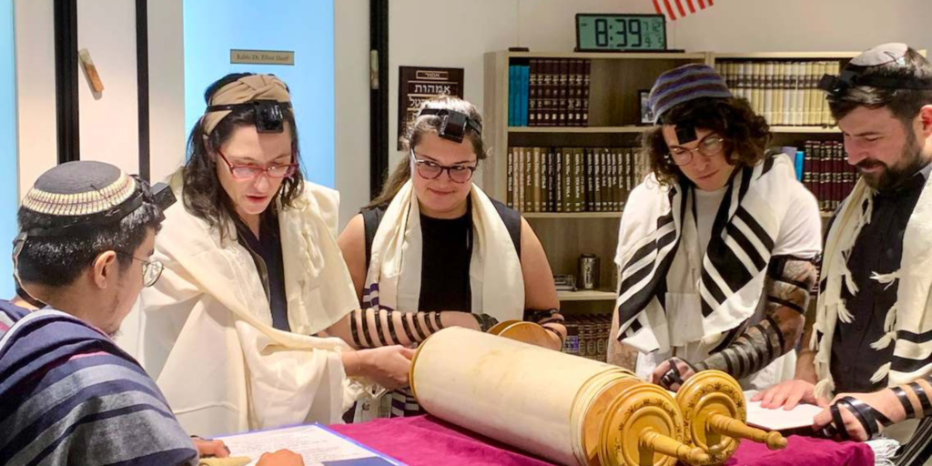 Five students wrapped in tefillin and talits stand over a closed torah in a room full of books as one student reads from a separate sheet of paper