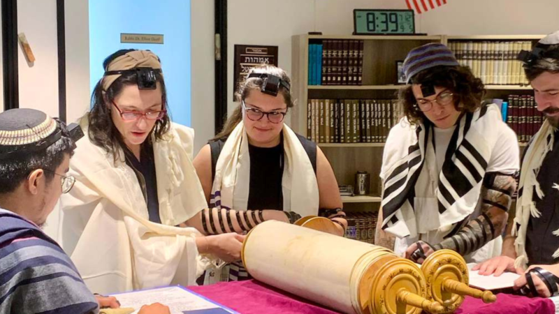 Five students wrapped in tefillin and talits stand over a closed torah in a room full of books as one student reads from a separate sheet of paper