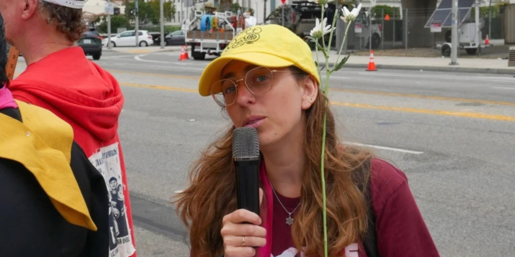 A young woman in a red shirt and Star of David necklace and yellow ball cap stand on the sidewalk against the street and talks into a hand mic
