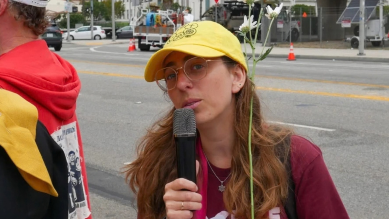 A young woman in a red shirt and Star of David necklace and yellow ball cap stand on the sidewalk against the street and talks into a hand mic