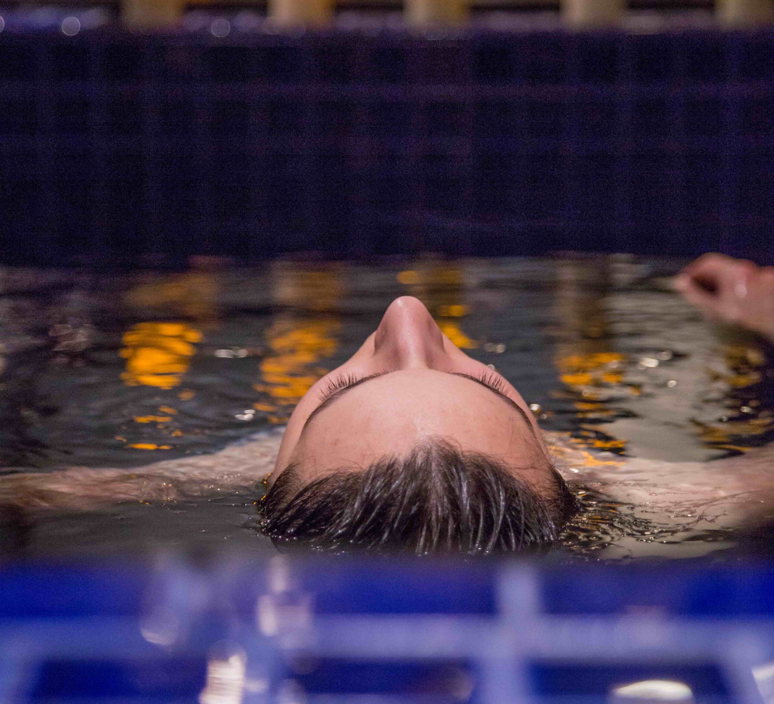 A person floating on their back in the mikvah bath with candles let above them on a shelf.